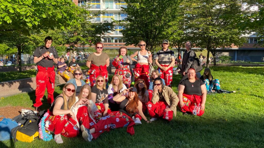 A photo of a group of people, most of whom are wearing red student overalls. All of them are smiling. The picture is taken in a park on a sunny day. In the background, more people in overalls of various colors can be seen sitting or standing.