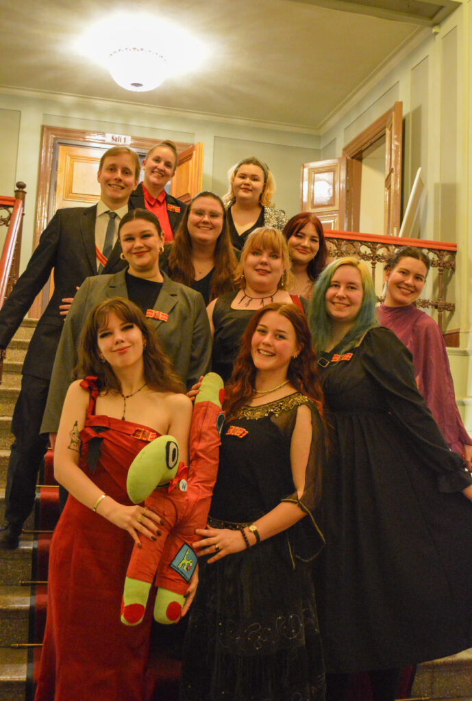 A photo of a group of 11 people standing indoors on a staircase. They are all smiling and dressed in either dresses or suits. Each person is wearing HUMAKO’s red-and-gold student union ribbon—either folded into a bow on the left side of the chest or worn diagonally from the right shoulder across the chest. The two people standing at the front are holding HUMAKO’s mascot, Hjördis: a green, H-shaped plush creature wearing red student overalls.