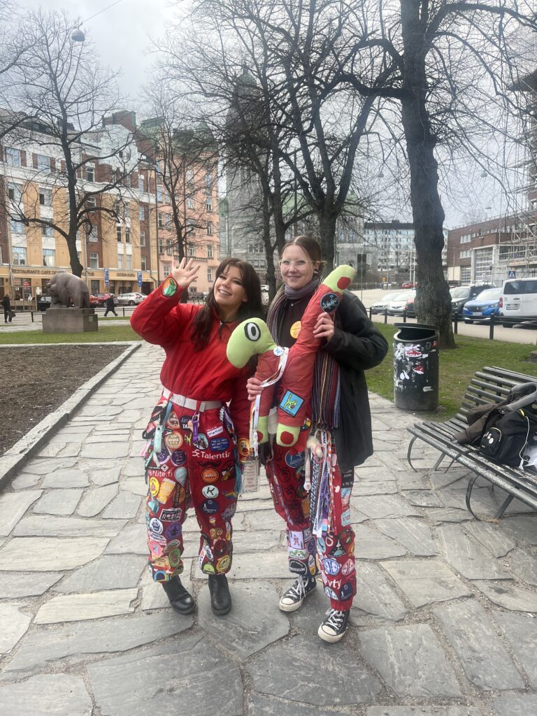 A photo of two smiling people wearing red student overalls. The person on the left has their hand slightly raised in a greeting gesture. The person on the right is holding Hjördis, HUMAKO’s mascot — a green, H-shaped plush creature dressed in red overalls. The photo is taken in a park; in the background, there are leafless trees, buildings, and a road with cars.