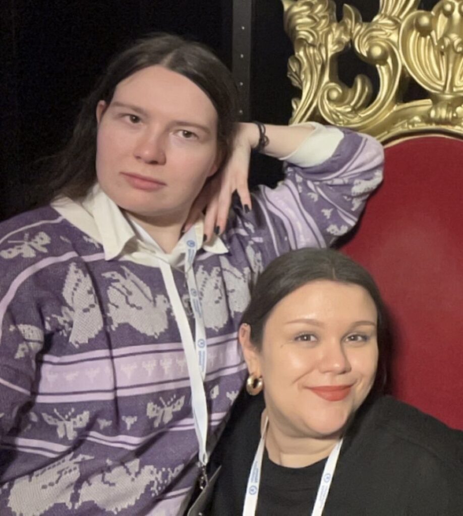 A photo of two people, both wearing lanyards around their necks. The person on the left has long hair, is wearing a purple top, and is leaning their elbow on the backrest of a chair while resting their hand against their cheek. The person on the right is smiling, has their hair tied back, and is sitting on a chair, appearing lower in height than the person next to them.
